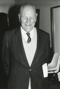 Older man in suit jacket, knit vest, white shirt and tie smiling towards camera
