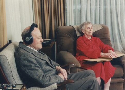 Jim wears headphones whilst Edith reads Braille in their lounge room