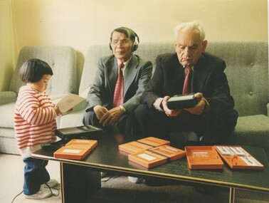 Young girl looks at booklet, whilst older man listens and second older man holds tape player