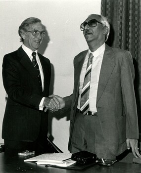 Two men shaking hands with a Braille book and cassette player on the table in front of them