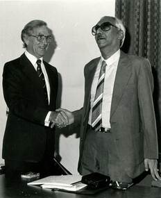 Two men shaking hands with a Braille book and cassette player on the table in front of them