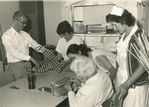Nursing staff and visitors look at handicrafts as women threads machine