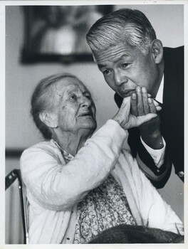 Japanese man leans over to hold hand of seated elderly woman and for her to touch his face