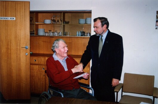 Man shaking hands with another in a wheelchair inside a conference room