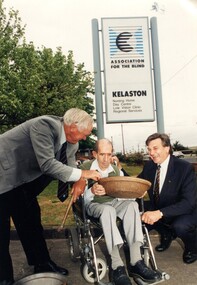 Two men smile as third man holds and looks into panning basin under Kelaston sign