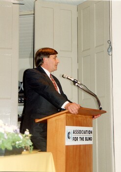 Man in suit at podium with table to his right and reel to reel player behind concertina doors