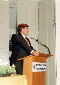 Man in suit at podium with table to his right and reel to reel player behind concertina doors