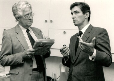 One man holds a newspaper and listens to another holding an small magnifier