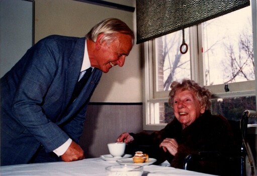 Older man speaking to seated elderly woman who has a teacup and cupcake in front of her