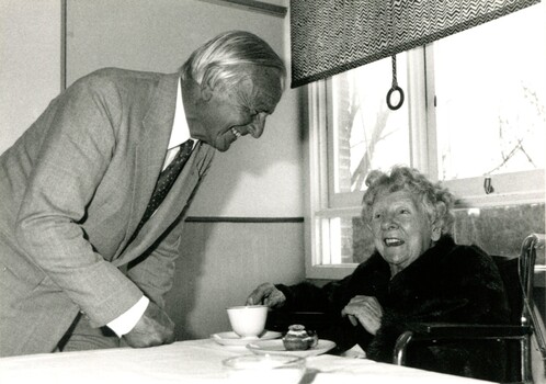 Older man speaking to seated elderly woman who has a teacup and cupcake in front of her
