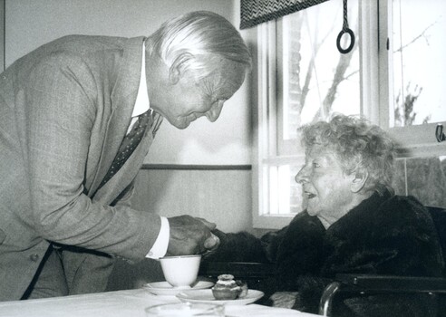 Older man speaking to seated elderly woman who has a teacup and cupcake in front of her
