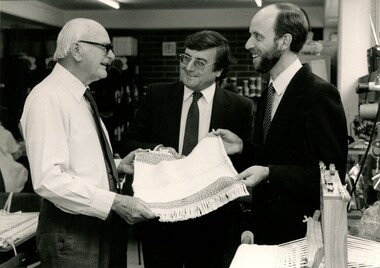 Three men hold a small pieces of weaving between them