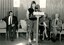 Speaker at podium whilst John Wicking and others look on