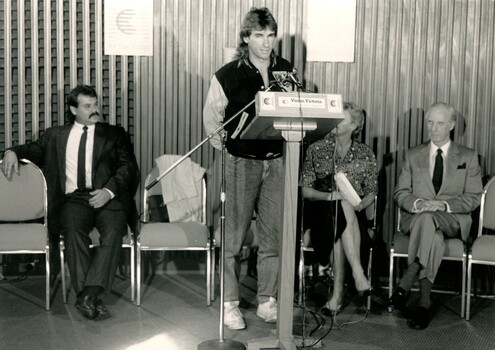Speaker at podium whilst John Wicking and others look on