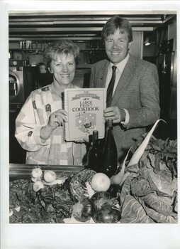 Female and male stand in kitchen holding copy of cookbook