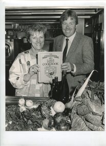 Female and male stand in kitchen holding copy of cookbook