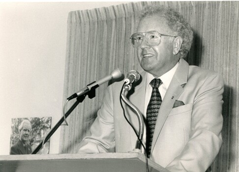 Man in suit and tie speaking to an audience with image of Arthur Wilkins beside him