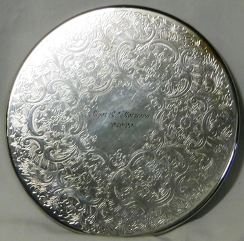 Circular silver place mat with engraving at centre.
