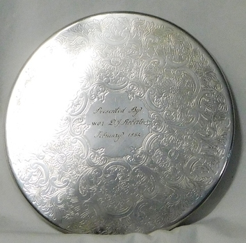 Circular silver place mat with engraving at centre.