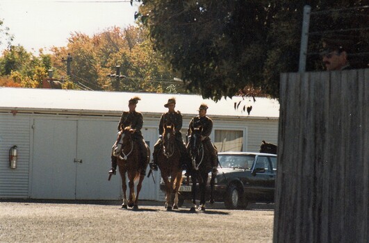 Three soldiers on horseback followed by a car near sheds.
