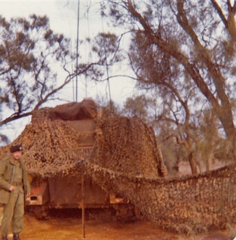 Soldier standing near armoured car covered with camouflaged netting in bushland.