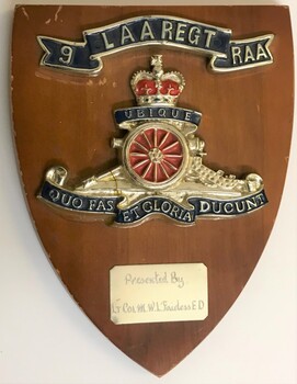 A wooden shield with a badge, title and small label attached to it.