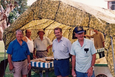 Five men standing outside camouflaged tent.