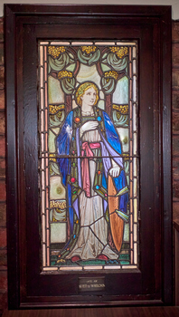 Coloured stained glass window showing the figure of Fortitude wearing a blue robe and holding a shield.  