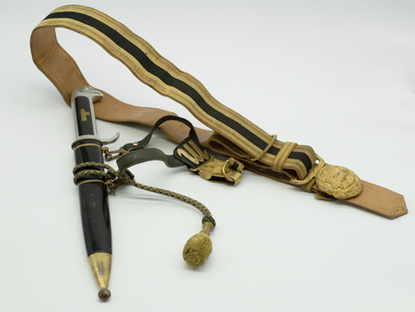 Black and gold scabbard with metal dagger, with gold and black belt with round gold coloured metal buckle. Gold fabric tassle hanging from the dagger