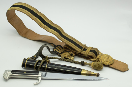 Metal dagger, silver handle with black inlay on grip, unsheathed from black scabbard with brass tip. Laid flat next to black and gold belt with gold coloured buckle, on white background.