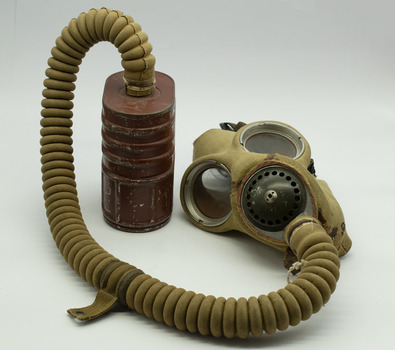 Khaki coloured mask with large glass eye pieces and metal respirator at nose, same fabric covered tube from base of mask to top of tin canister. Canister is red/brown colour oval shape, on white background.