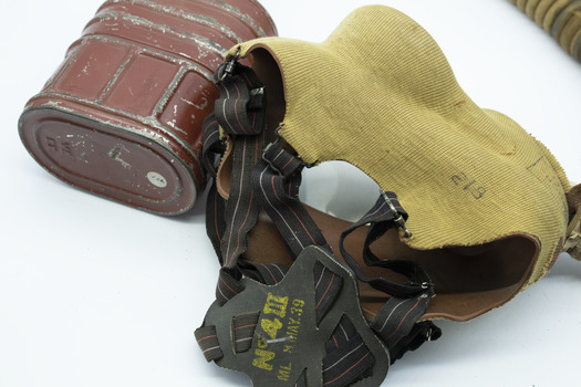 Khaki covered mask with black rubber head straps, same fabric covered tube from base of mask to top of tin canister. Canister is red/brown colour oval shape, lying with base visible on white background.