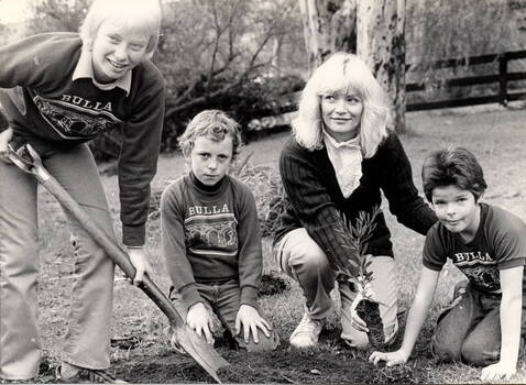 Three children supervised by an adult are planting a tree.