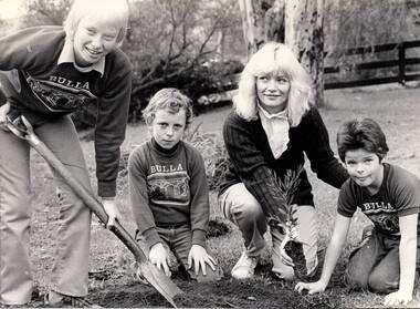Three children supervised by an adult are planting a tree.