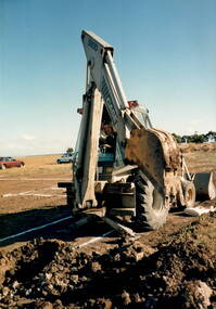 A coloured photograph of a lady driving a large grader in an open area.