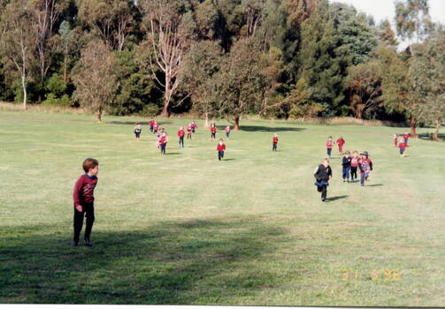 A number of school children running across an open cleared area with trees growing in the background.