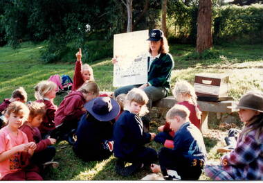 A lady is seated in front of a group of children in an outdoor area. She is holding up a chart showing various insects.