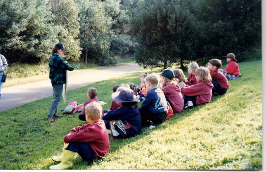 Children are seated on a grassy hillside while listening to a teacher who is holding up a chart and explaining it to the group.