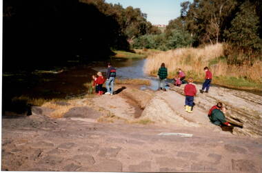 Eight children with two adults are standing on a rocky outcrop beside a river and looking for the life that is in the water.
