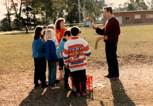 A group of school children are listening too a man holding two boomerangs.