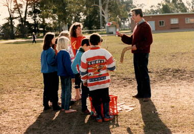 A group of school children are listening too a man holding two boomerangs.