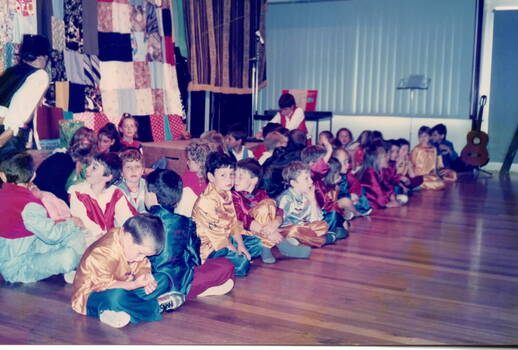 Children in fancy dress seated on the floor in a hall.