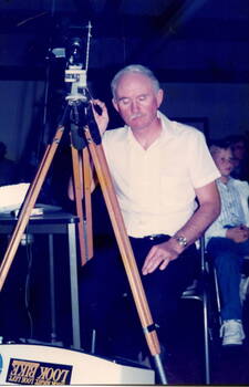 A man standing behind a camera which is mounted on a tripod.