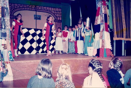 Ten children in fancy costume from the upper primary classes are performing on the stage with two older girls leading the group.