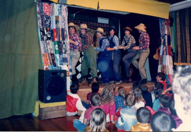 Children are watching six ladies dressed as bushmen dancing on a stage in a hall.