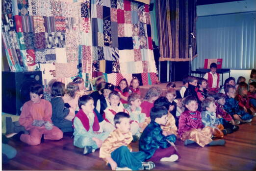 A number of children, dressed in fancy dress, are seated in front of a stage on a wooden floor.