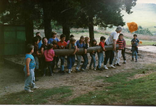 Seventeen children and an adult carrying along log in an open area.