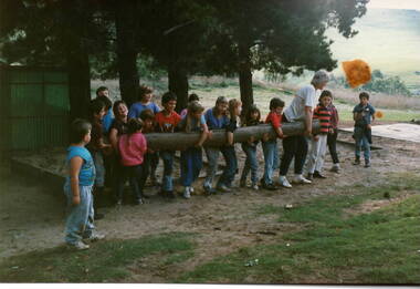 Seventeen children and an adult carrying along log in an open area.