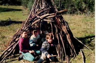 Three children sitting at the entrance of a bark shelter.