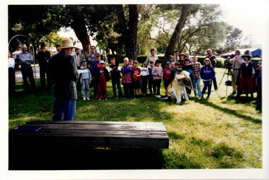A group of children and teachers standing in a shaded area watching a man talking to them.